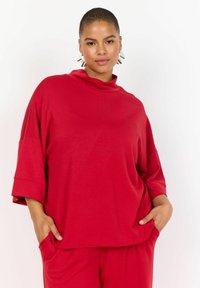 Červený oversized top s vysokým výstrihom, spadnutými ramenami a krátkymi rukávmi. Vyrobený z mäkkej, textúrovanej látky, má bočné vrecká.