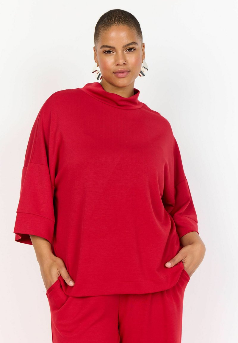 Červený oversized top s vysokým výstrihom, spadnutými ramenami a krátkymi rukávmi. Vyrobený z mäkkej, textúrovanej látky, má bočné vrecká.