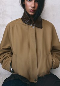 Ženska s temnimi lasmi, oblečena v bež jakno z temno rjavim usnjenim ovratnikom, z rokami v žepih, ob svetlo sivi teksturirani steni.