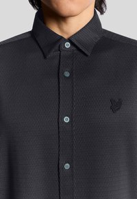 Polo shirt nera con tessuto testurizzato, colletto a polo abbottonato, bottoni blu e un piccolo logo nero sul lato sinistro del petto.