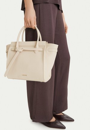 Osoba držící béžovou koženou kabelku s horními uchy a odnímatelným popruhem, oblečená v tmavých širokých kalhotách a černých špičatých botách.