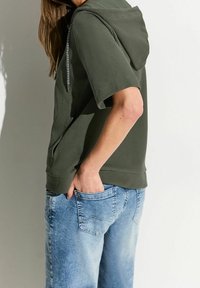 Green hooded sweatshirt with short sleeves and a textured finish. Paired with light blue denim jeans, visible pocket detail.