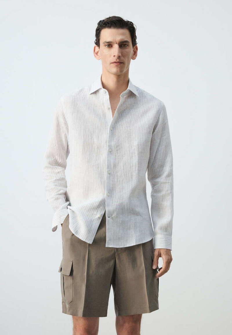Homme portant une chemise blanche rayée à manches longues boutonnée et un short cargo kaki, debout devant un fond uni.