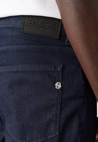 Tamni traperice s teksturiranim kožnim pojasom na kojem je ugravirano "BOSS", srebrni detalj na stražnjem džepu, pojednostavljen dizajn.