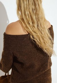 Maglione marrone a spalle scoperte in maglia con una finitura texturizzata. Lunghi capelli biondi ondulati parzialmente visibili, mentre tiene una borsa marrone.