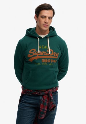 Mann trägt grünen Hoodie mit "SuperDry"-Logo, karierte Hemd um die Taille gebunden und blaue Jeans, steht mit den Händen hinter dem Rücken.