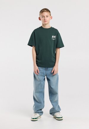 Adolescent garçon debout portant un t-shirt NYC vert foncé, un jean bleu clair ample et des baskets blanches sur un fond blanc uni.