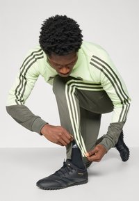 Athletic outfit featuring a light green long-sleeve top with black stripes and olive green pants. Black soccer shoes with yellow accents.