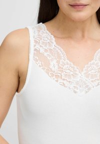 Bílý žebrovaný tílko s květinovým krajkovým lemem kolem krku, s jemnou strukturou a přiléhavým střihem. Žádné další detaily ani vzory nejsou viditelné.