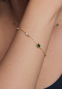 Złoty bransoletka łańcuszkowa z naprzemiennie umieszczonymi zielonymi i przezroczystymi kamieniami szlachetnymi. Projekt jest delikatny, z misternie wykonanym łańcuszkiem i pewnymi mocowaniami.