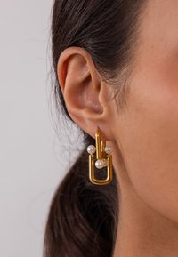 Ohrringe in Goldfarbe mit minimalistischer Gestaltung, die eine U-förmige Struktur und drei weiße Perlen zeigen. Glatte Textur und modernes ästhetisches Design.