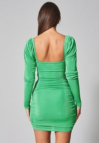 Robe longue verte à manches longues avec un dos carré, fabriquée en matériau extensible, présentant des fronces sur les côtés pour une silhouette ajustée.
