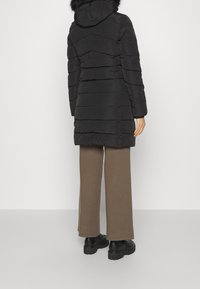 Abrigo tipo puffer negro con diseño acolchado horizontal y capucha de piel sintética. Combinado con pantalones marrones de pierna ancha y botas negras robustas.