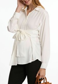 Weißes Bluse mit gebundenem Saum, Kragen und Knopfleiste. Lange Ärmel mit Knopfmanschetten, aus glattem, fließendem Stoff.