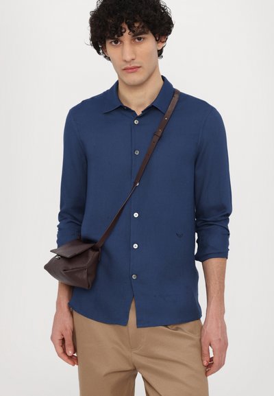 Navyblauw hemd met lange mouwen, knoopsluiting en opgerolde mouwen, gecombineerd met een bruin leren schoudertas, op een neutrale achtergrond.