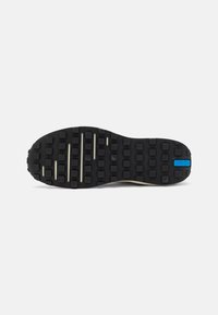 Black rubber sole with raised squares and grooves, featuring a blue accent and cream-colored sections for texture contrast.