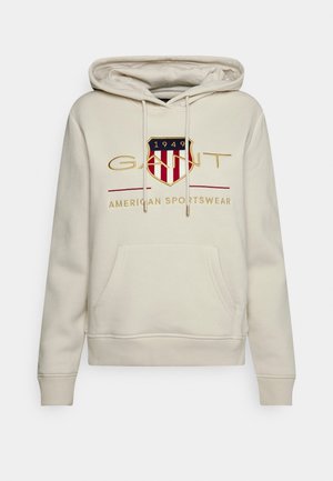 Sudadera con capucha color crema con bolsillo frontal, cordones y logotipo bordado en forma de escudo que dice "1949 GANT American Sportswear" en el pecho.