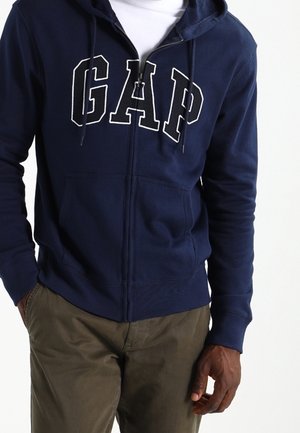 Sudadera con cremallera de color azul marino, hecha de algodón, que presenta un logotipo blanco de "GAP" en el frente, con capucha ajustable por cordón y bolsillos frontales.