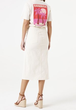 Spódnica dżinsowa w kolorze kremowym z dopasowaną talią i tylnymi kieszeniami. Noszona z beżowym t-shirtem z nadrukiem przedstawiającym wschód słońca i palmy.