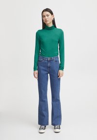 Grüner langärmeliger Rollkragenpullover, kombiniert mit blauen Schlagjeans und schwarzen Turnschuhen. Das Outfit zeichnet sich durch ein tailliertes Design und eine weiche Textur aus.