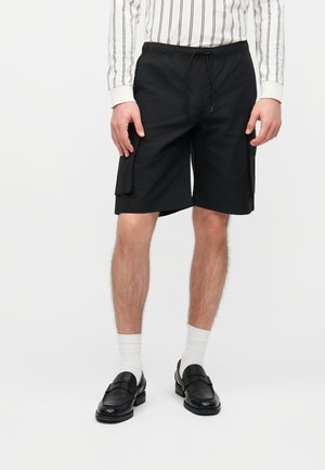 Muž nosi čierne cargo šortky s bočnými vreckami, biele ponožky, čierne mokasíny a bielu košeľu s dlhým rukávom s čiernymi vertikálnymi pruhmi.