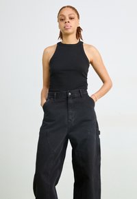 Svart ribbet tanktop kombinert med løstsittende svarte denimbukser med sidelommer og høyt liv. Enkelt bakgrunn.