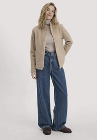 Beige Zip-Jacke mit Reißverschluss, hochgeschlossenes cremefarbenes Oberteil, weite blaue Jeans und dunkelbraune Schuhe, vor einem schlichten hellen Hintergrund.