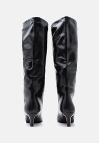 Μαύρες μπότες μέχρι το γόνατο από γυαλιστερό υλικό, με λεπτό τακούνι, λεία υφή και ρυτιδωτή λεπτομέρεια κατά μήκος του κορμού.