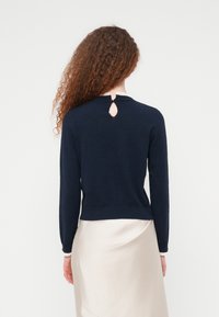 Άτομο με μακριά σγουρά μαλλιά που φοράει μπλε μπλούζα με μακριά μανίκια και κλείσιμο με κουμπί στο λαιμό και ανοιχτό μπεζ φούστα, εμφανίζεται από πίσω.