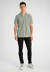 Πικέ πράσινο πόλο μπλουζάκι με μαύρες κατακόρυφες ρίγες, αναδιπλούμενος γιακάς και άνετη εφαρμογή, συνδυασμένο με μαύρο παντελόνι και άσπρα αθλητικά παπούτσια.