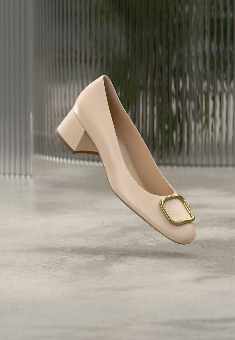 Beež madala kontsaga naisteking ruudukujulise kuldse pandlaga varba kohal, taustal sile betoonpõrand ja vertikaalselt triibuline klaaspind.