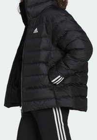 Veste noire matelassée avec un design matelassé, un col montant, une fermeture éclair et trois bandes blanches sur le poignet. Logo Adidas bien visible sur la poitrine.