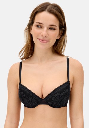 Mujer de cabello castaño claro con un sujetador push-up de encaje negro, sonriendo ligeramente y mirando hacia su izquierda sobre un fondo liso.