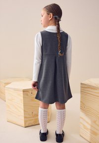 Ragazza con lunghi capelli intrecciati, che indossa un vestito grigio, una camicetta bianca, calzini a motivi fino al ginocchio e scarpe nere, in piedi tra casse di legno.