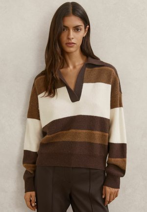 Vrouw draagt een bruin, wit en beige breisel met brede strepen, een diepe V-hals en donkerbruine broek tegen een neutrale achtergrond.