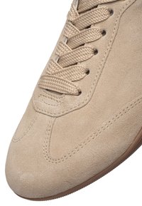 Detailní záběr na špičku béžové semišové tenisky s odpovídajícími tkanými tkaničkami a detailním šitím podél okrajů a podrážky.