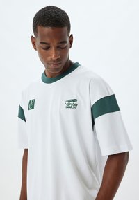 Camiseta de algodón blanca con detalles en verde en las mangas y el cuello, con logo impreso en el pecho. Textura suave, corte relajado.