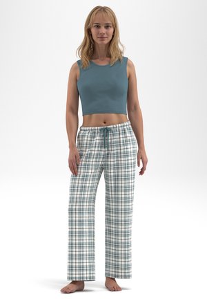 Young woman standing barefoot wearing a sleeveless blue crop top and white and blue plaid pajama pants against a plain background.