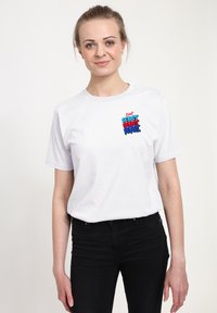 Bílý bavlněný tričko s barevným, texturovaným grafickým motivem "Pij!" s červenými, modrými a černými písmeny na hrudi. Krátké rukávy, kulatý výstřih.