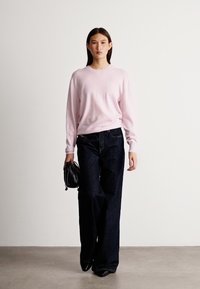 Ανοιχτό ροζ πλεκτό πουλόβερ συνδυασμένο με φαρδιά σκούρα μπλε τζιν και κρατώντας μια μικρή μαύρη τσάντα, σε ουδέτερο φόντο.