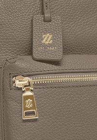 Sección de bolsa de cuero gris con cierre de cremallera dorada y etiqueta de marca con "ZZ" y "EST 2007" en relieve. Superficie texturizada con grano visible.