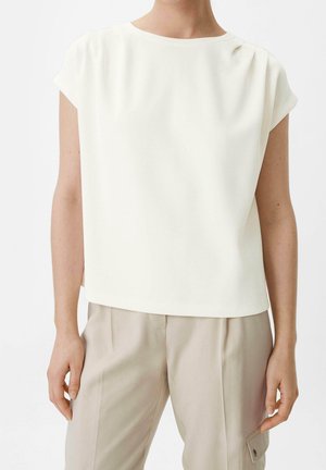 Person iført en løs, kortærmet hvid top med rund hals og lyse beige bukser med en sidelommeklap, stående mod en ensfarvet baggrund.