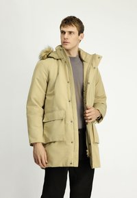 Hombre vestido con un abrigo de invierno beige con capucha de piel, suéter gris y pantalones negros, mirando hacia un lado sobre un fondo sencillo.