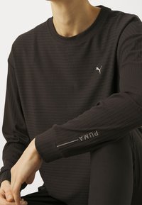 Schwarzes Langarmshirt aus geripptem Stoff, mit einem kleinen weißen Puma-Logo auf der Brust und "PUMA" auf dem Bündchen bedruckt.