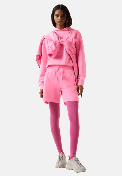 Femme portant un sweat à capuche rose vif, un short assorti rose, des collants roses et des baskets beige, debout devant un fond blanc.