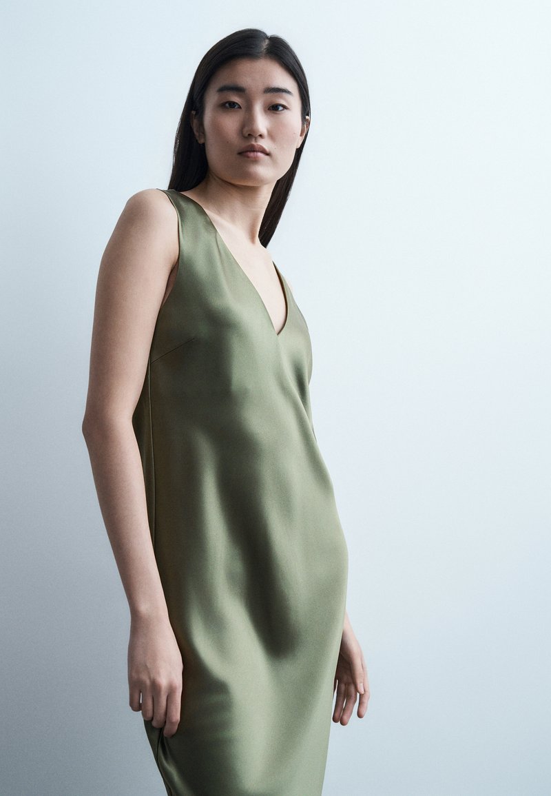 Νεαρή γυναίκα με μακριά μαύρα μαλλιά, φορώντας αμάνικο σατέν φόρεμα σε ελιά πράσινο χρώμα, μπροστά σε απλό ανοιχτό φόντο.