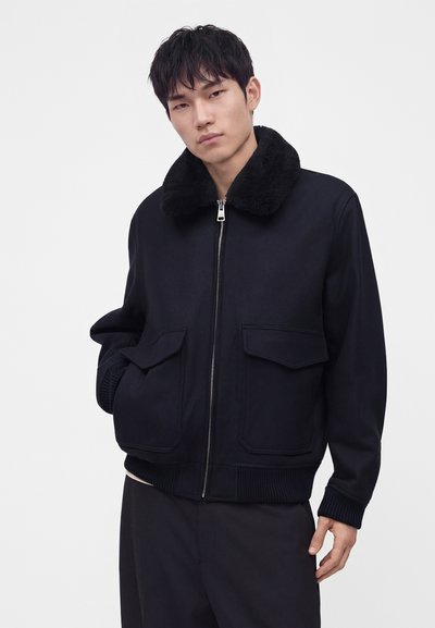 Jeune homme portant une veste bomber noire avec un col en fourrure épaisse, fermeture éclair à l'avant et poches à rabat, posant avec une main dans la poche.