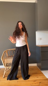 Top branco, sem mangas, com pontos decorativos, combinado com calças pretas largas. Cabelo encaracolado, chão de madeira e uma cadeira de vime ao fundo.