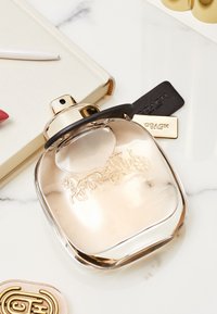 Klart glassparfymeflaske med avrundede kanter, gullaccent og svart hette. Har gravert Coach-logo og står på en marmoroverflate.