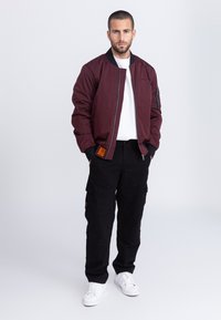 Man met kort haar en baard, gekleed in een bordeaux bomberjack, wit overhemd, zwarte cargobroek en witte sneakers, staand tegen een witte achtergrond.